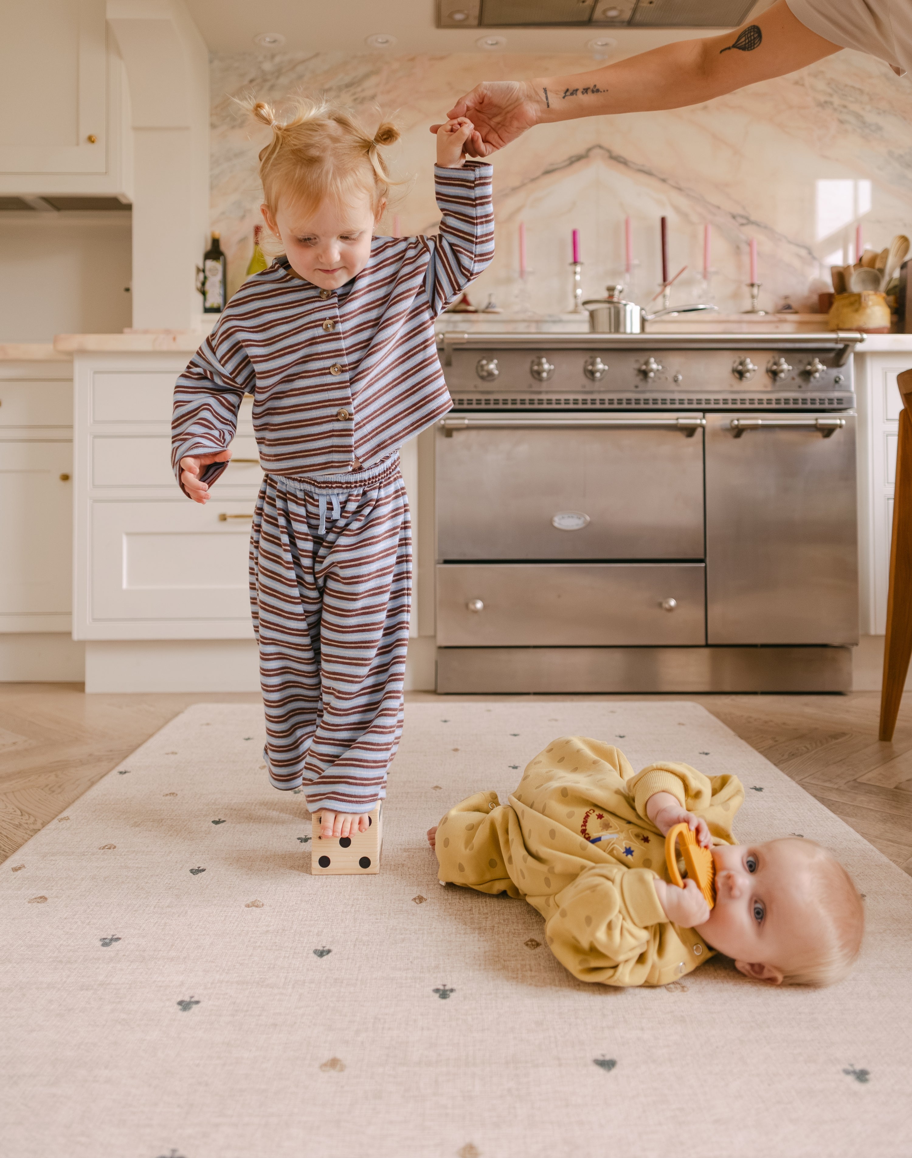 Toddler balancing in the kitchen on a The Wildcard Totter + Tumble reversible wipeable memory foam playmat while the baby lies on it